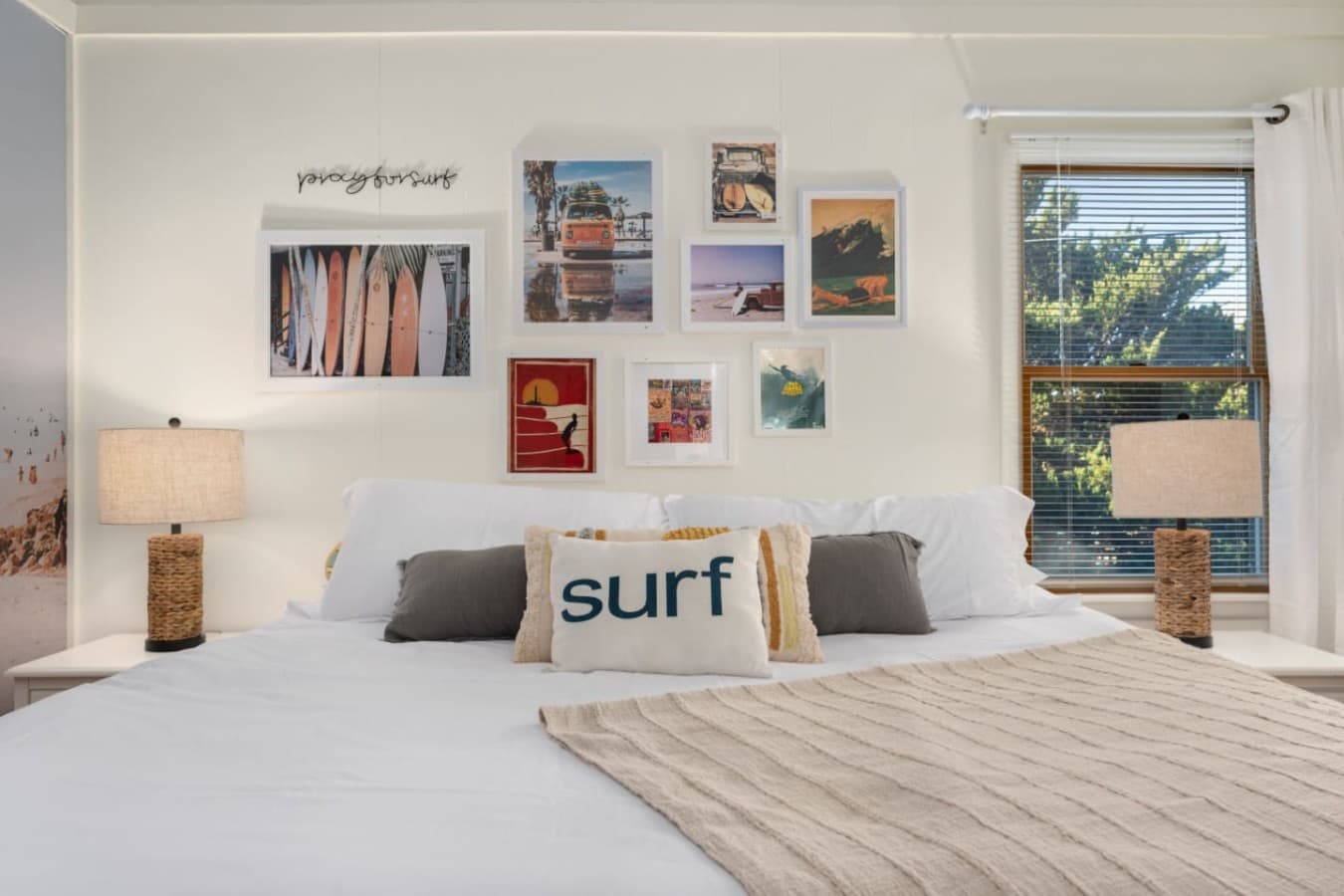 Relaxed bedroom w/ soft bedding, surf-themed pillows & cheerful natural light.