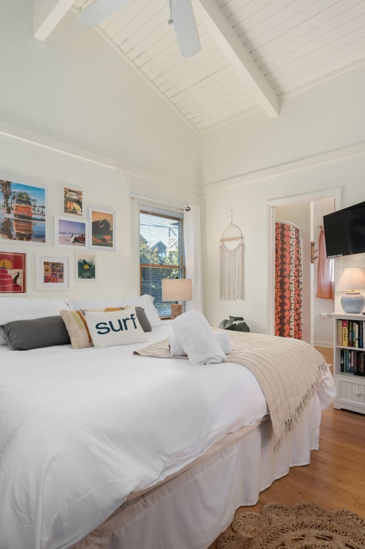 Bedroom w/ vaulted beamed ceiling, white bedding & a surf-themed gallery wall.
