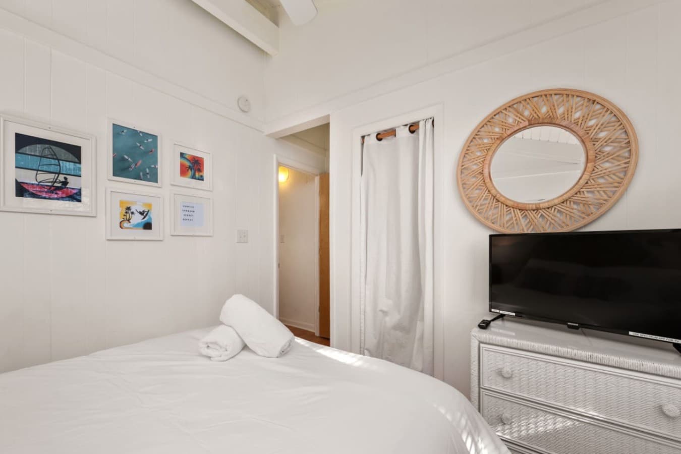 Bedroom w/ a TV on the dresser, rattan mirror & framed beach prints above the bed.