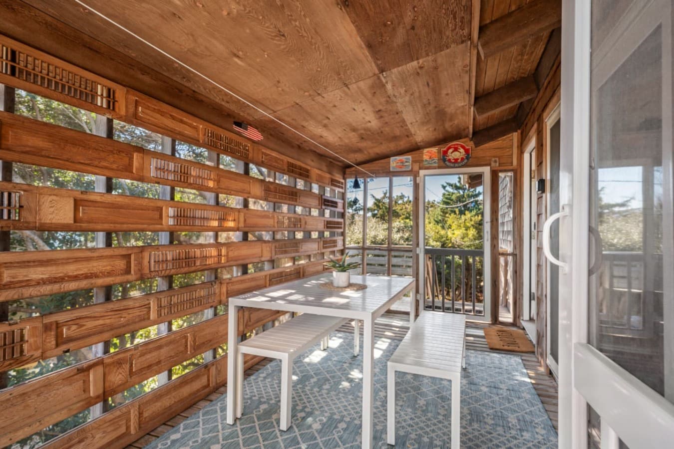 Shaded ground-level dining space w/ slatted wood walls, a long table & sandy floor.