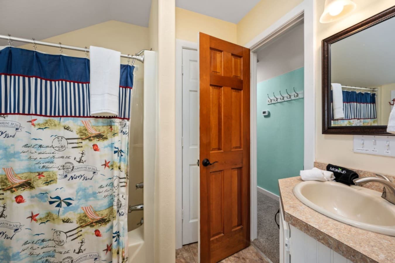 Bathroom w/ tub-shower behind a nautical curtain, vanity mirror & a row of towel hooks.