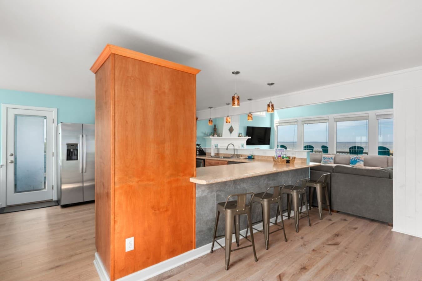 Kitchen bar w/ metal stools & a stainless fridge, open to the living room's water-facing windows.