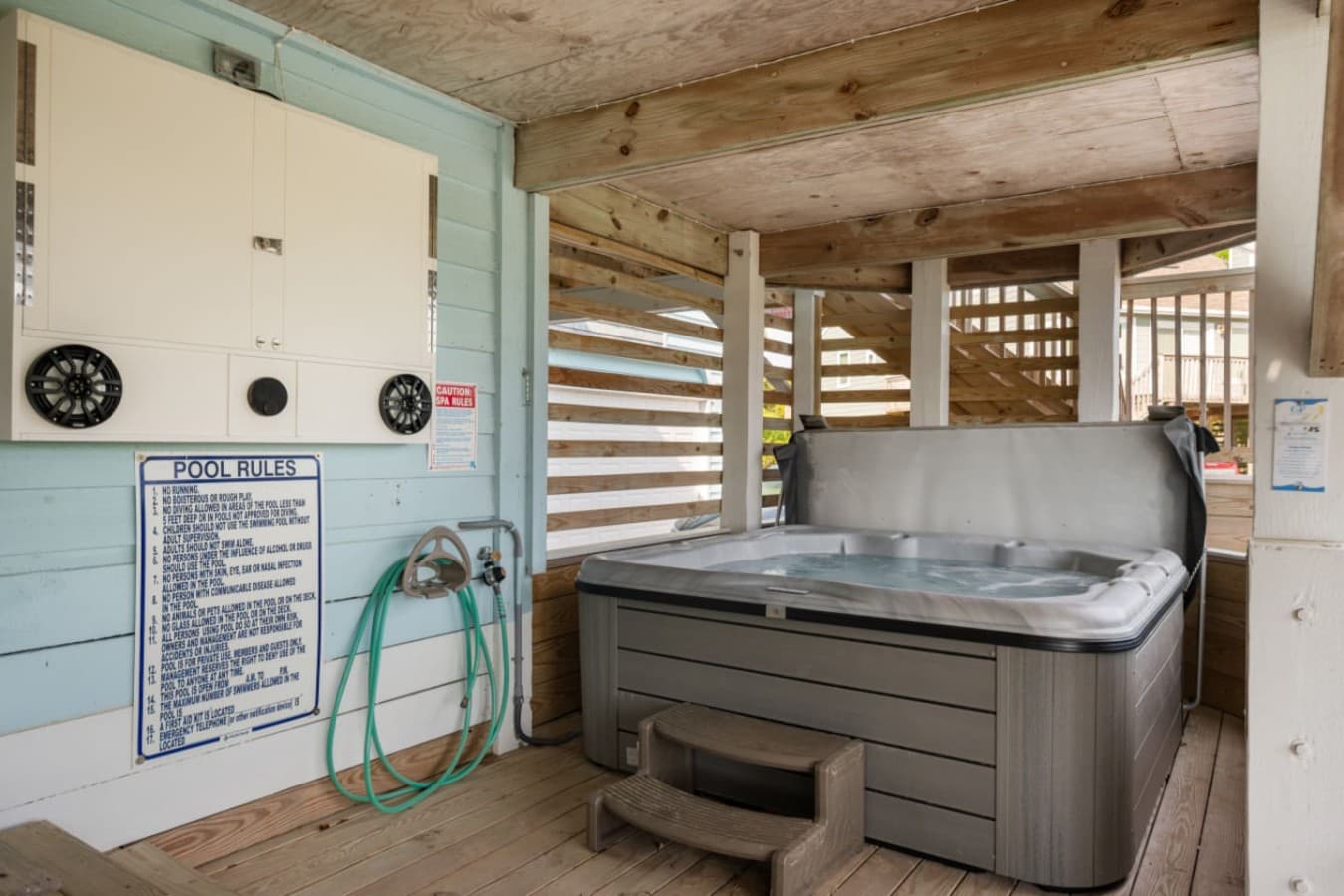 Covered hot tub on the wood deck w/ a rinse hose & posted pool rules alongside.