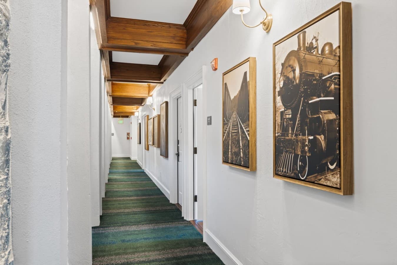 A clean and well-lit shared hallway leading to guest rooms featuring classic artwork and warm wood beam details.
