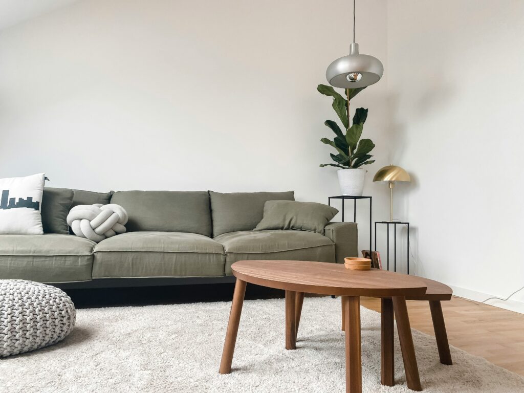 A living room staged with eco friendly furniture including a sage green sofa, neutral beige rug, and a set of medium stain wood coffee tables.